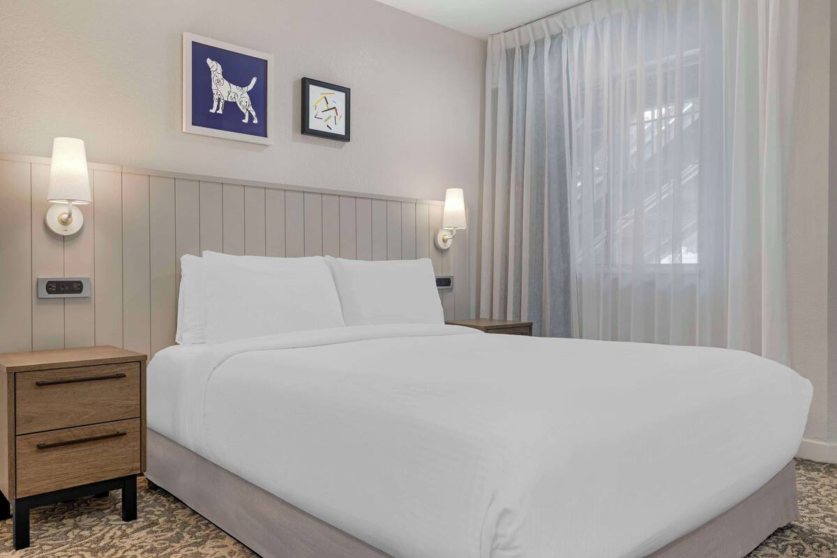 Ein ordentlich bezogenes Bett mit weißen Leinentüchern in einem Hotelzimmer. An beiden Seiten des Bettes befinden sich zwei Wandlampen, und über dem Bett hängt ein Bild von einem Hund.