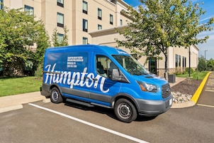 Una camioneta azul con la marca 'Hampton Inn by Hilton' estacionada frente a un edificio con múltiples ventanas.