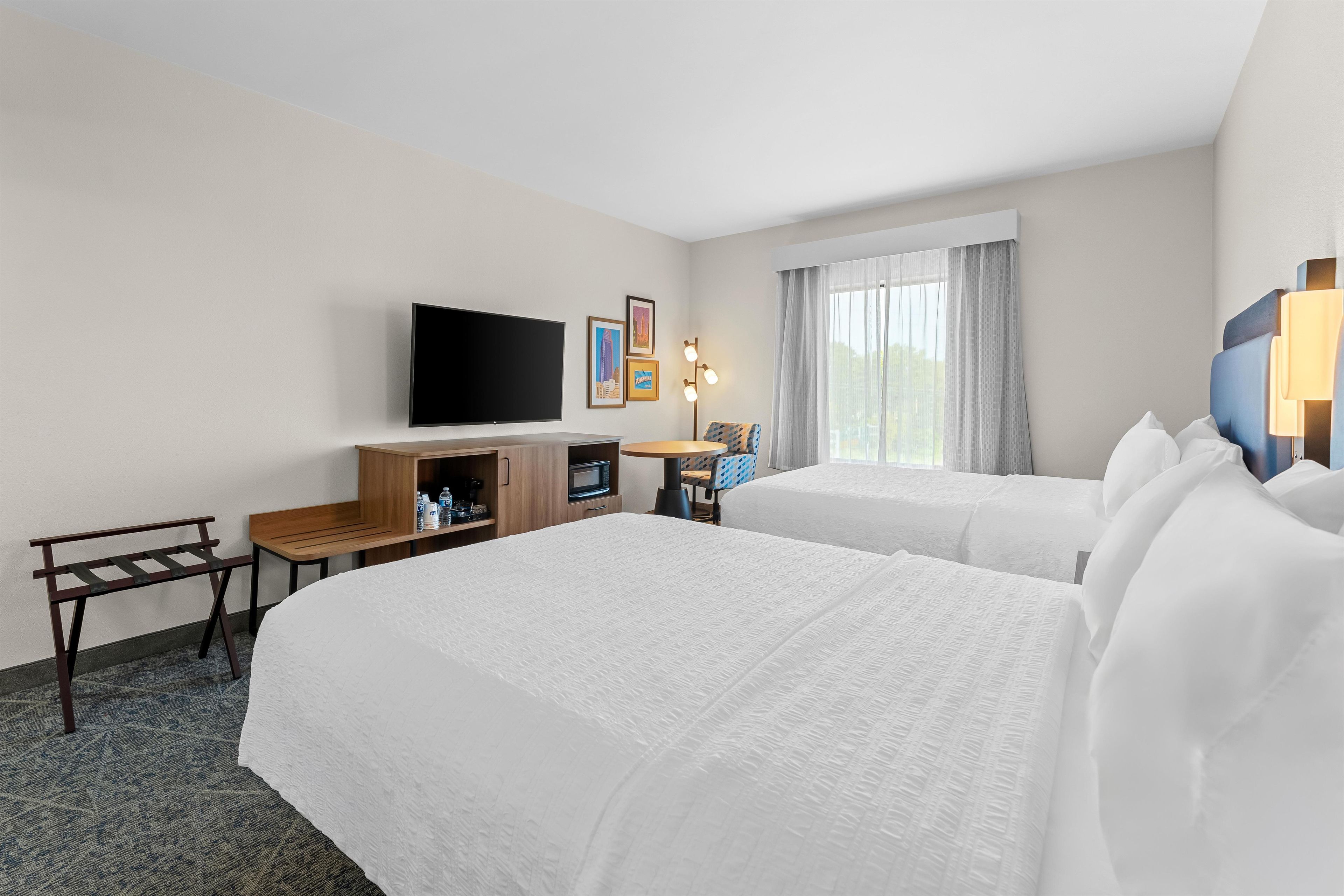 Habitación de hotel con dos camas, televisión, escritorio con silla, una mesita y ventana con cortinas.