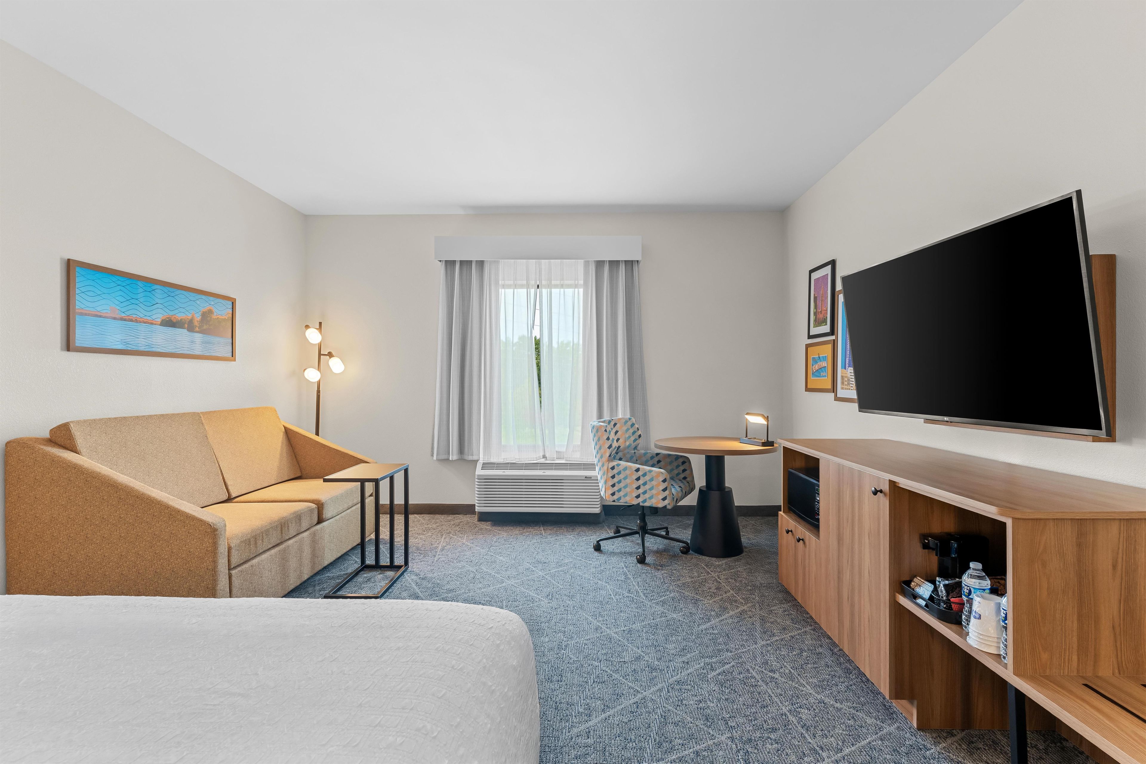 Habitación de hotel con sofá, una silla, televisor, escritorio y un amplio ventanal con cortinas.