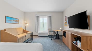 Habitación de hotel con sofá, una silla, televisor, escritorio y un amplio ventanal con cortinas.
