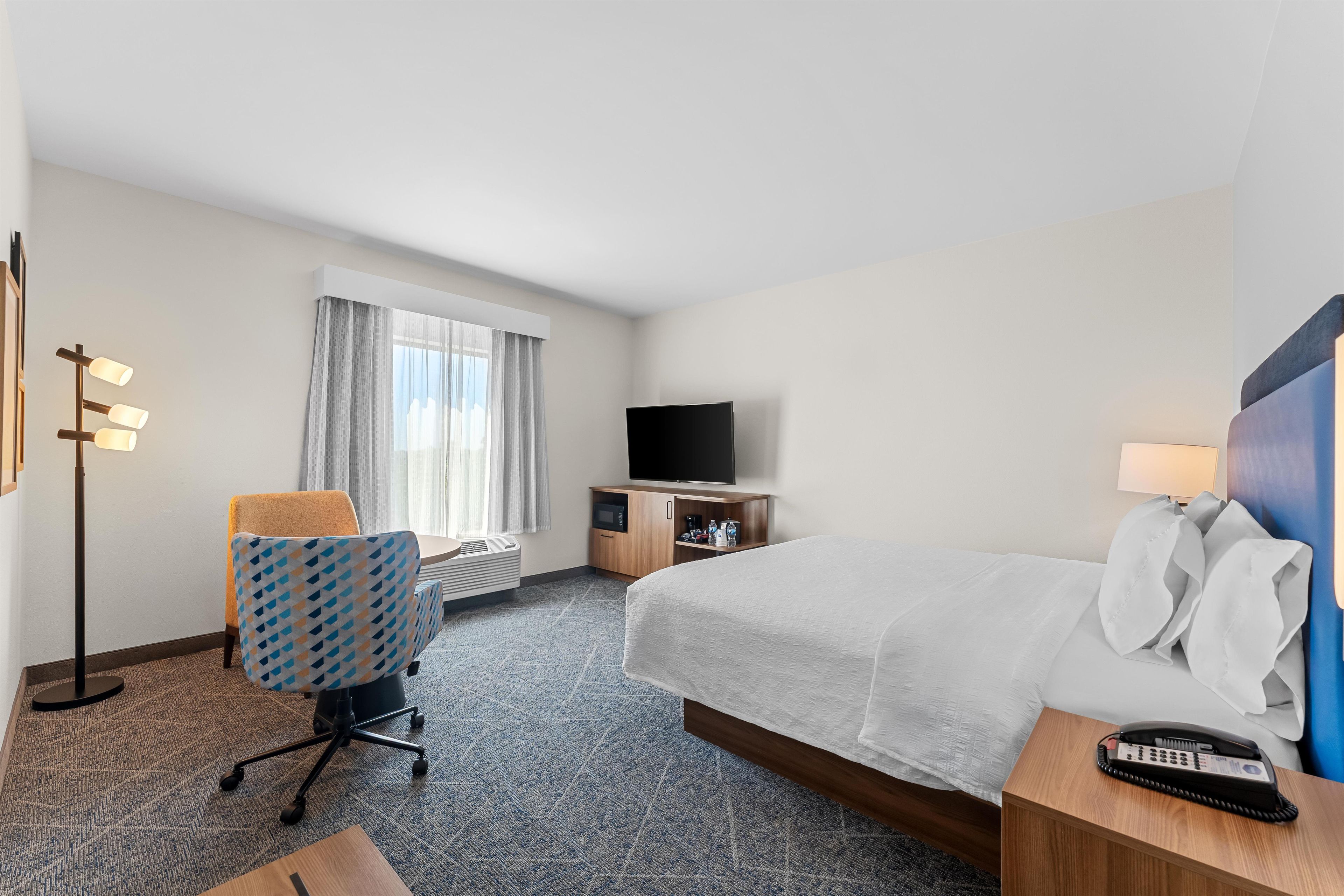 A hotel room with a bed, a desk, a chair, a television, and a bedside table with a phone.