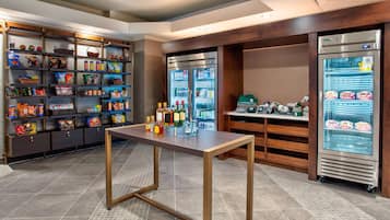 Estación autoservicio de aperitivos y bebidas con estantes llenos de diferentes opciones y un refrigerador con bebidas.