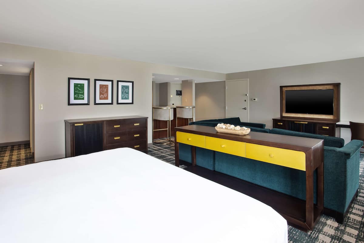 Habitación de hotel con una cama grande, un aparador de madera con cajones amarillos, un sofá color turquesa, un televisor de pantalla plana y obras de arte enmarcadas en la pared.