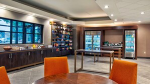A modern kitchen with a large window, built-in shelves with snacks, a refrigerator, and a dining area with orange chairs. - DoubleTree by Hilton Lansing (Lansing)