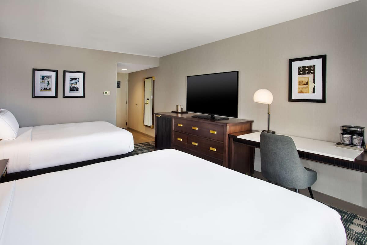Habitación de hotel con dos camas, un escritorio, una silla, un televisor de pantalla plana y cuadros en las paredes.