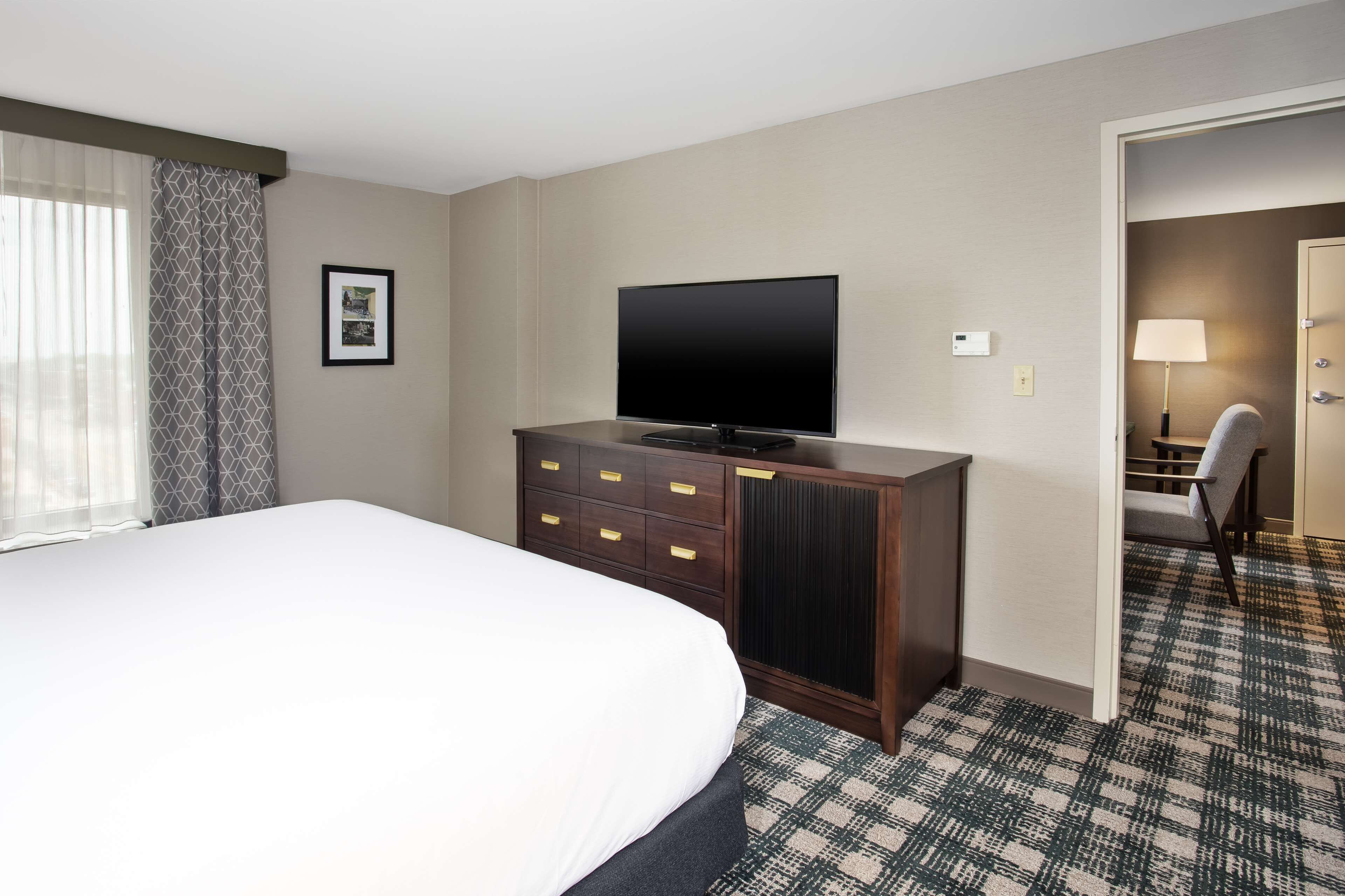 A hotel room with a large bed, a TV on a wooden dresser, a patterned carpet, and a door leading to another room.