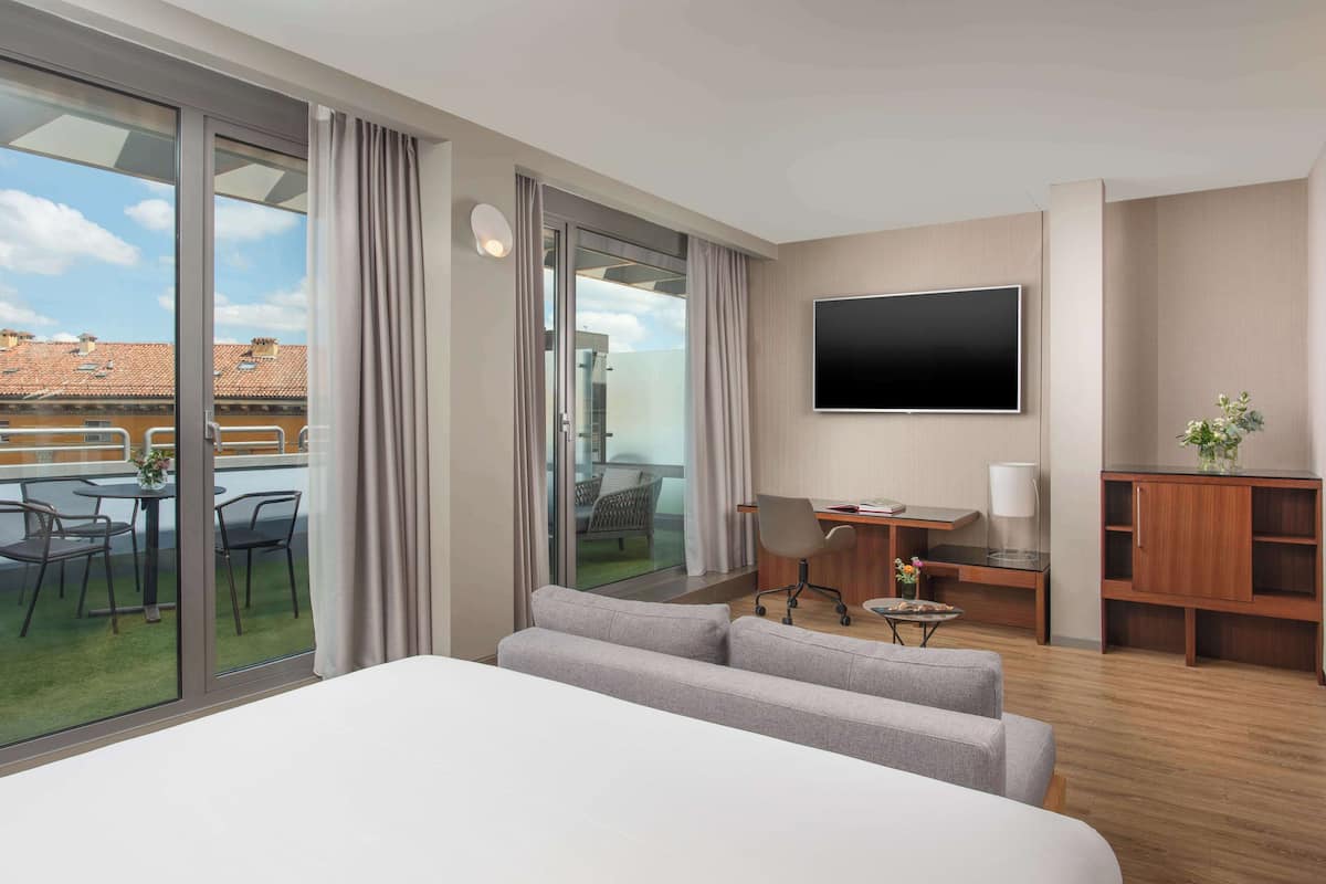A hotel room with a large bed, a desk with a chair, a TV, and a balcony with outdoor seating.