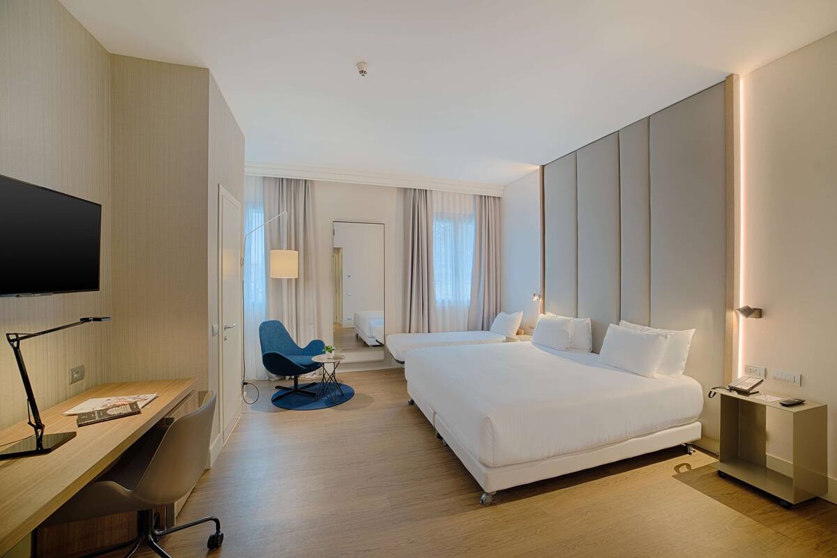A modern hotel room with a large bed, a desk with a chair, a television, and a bedside table with a lamp.