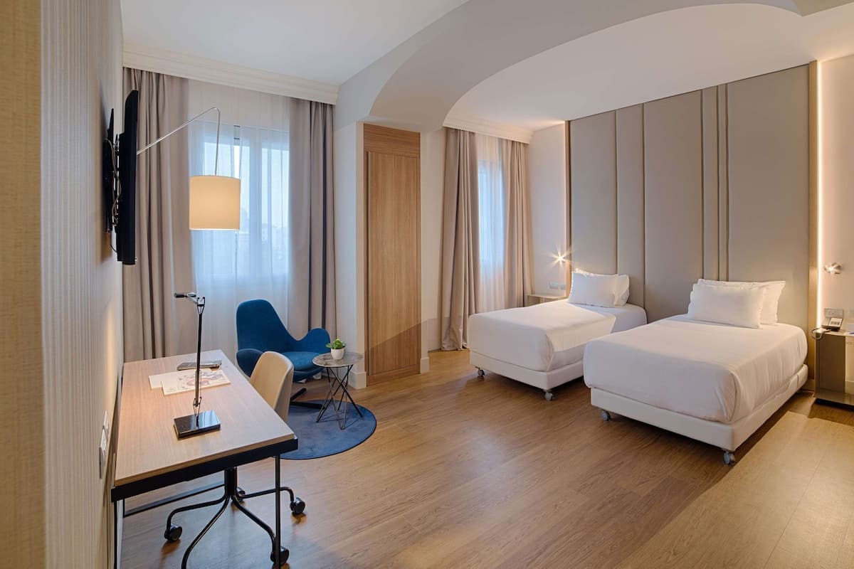 A hotel room with two beds, a desk, a chair, a lamp, and a TV.