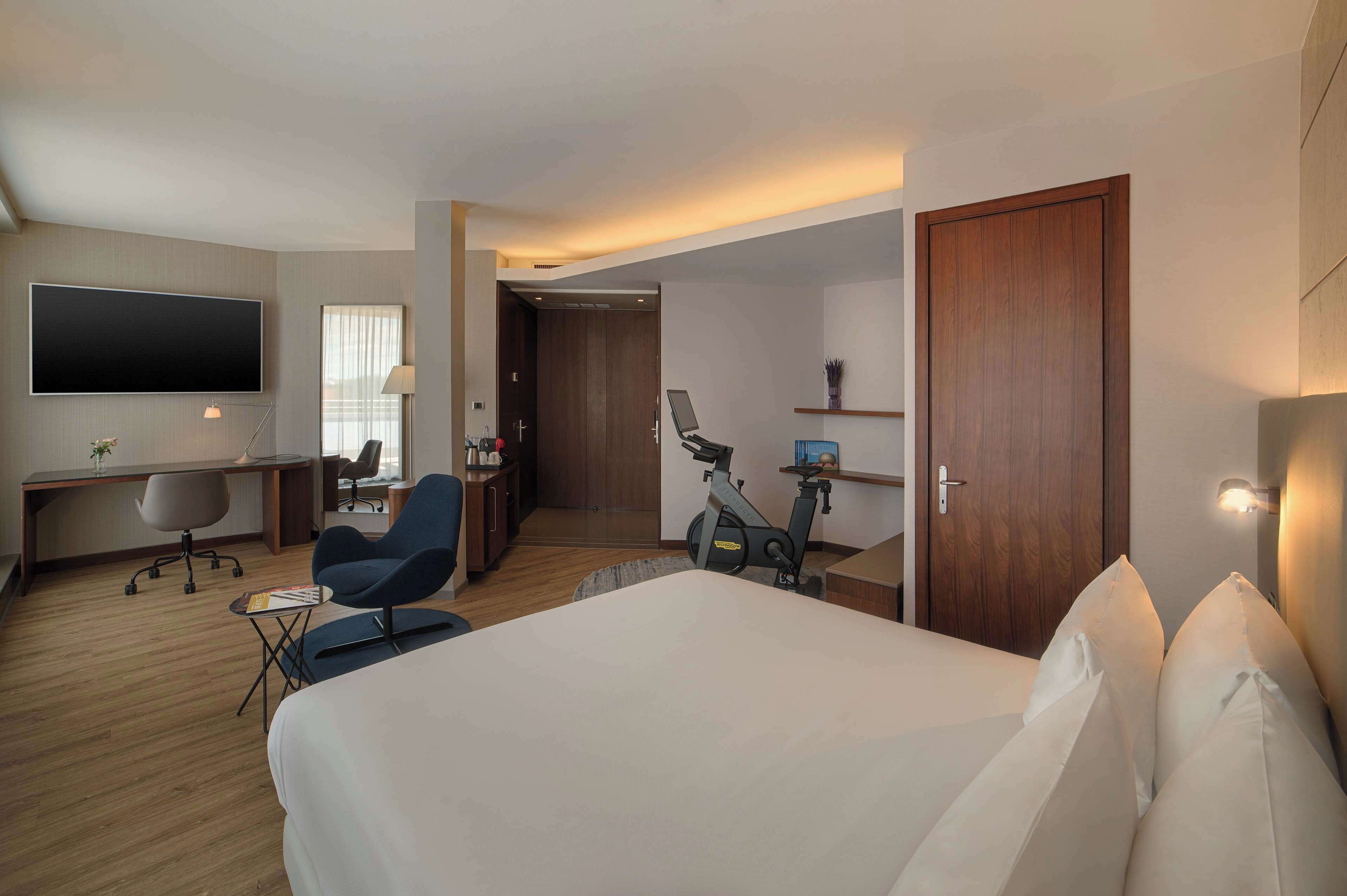 A hotel room with a large bed, a desk with a chair, a treadmill, and a TV mounted on the wall.