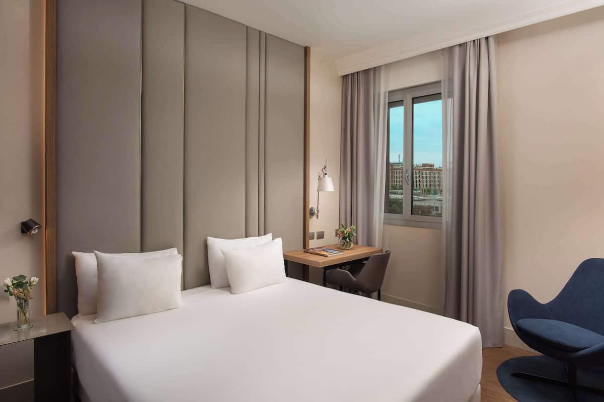 A hotel room with a large bed, a desk, and a chair. There is a window with curtains, a lamp, and a vase with flowers on the desk.