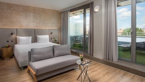 Habitación de hotel moderna con una cama grande, un sofá gris, una mesita con aperitivos y un balcón con vistas.