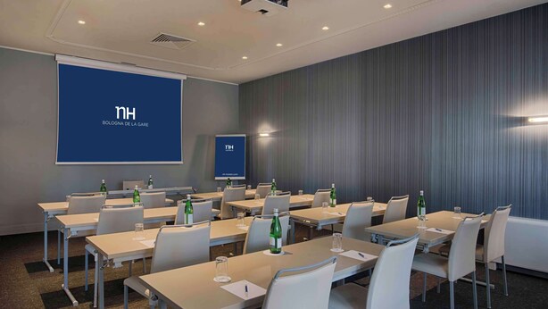 A conference room with multiple tables and chairs, a projector screen displaying 'nh Bolognà de la Gàre', and a smaller screen with the same logo. - NH Bologna de la Gare (Bologna)