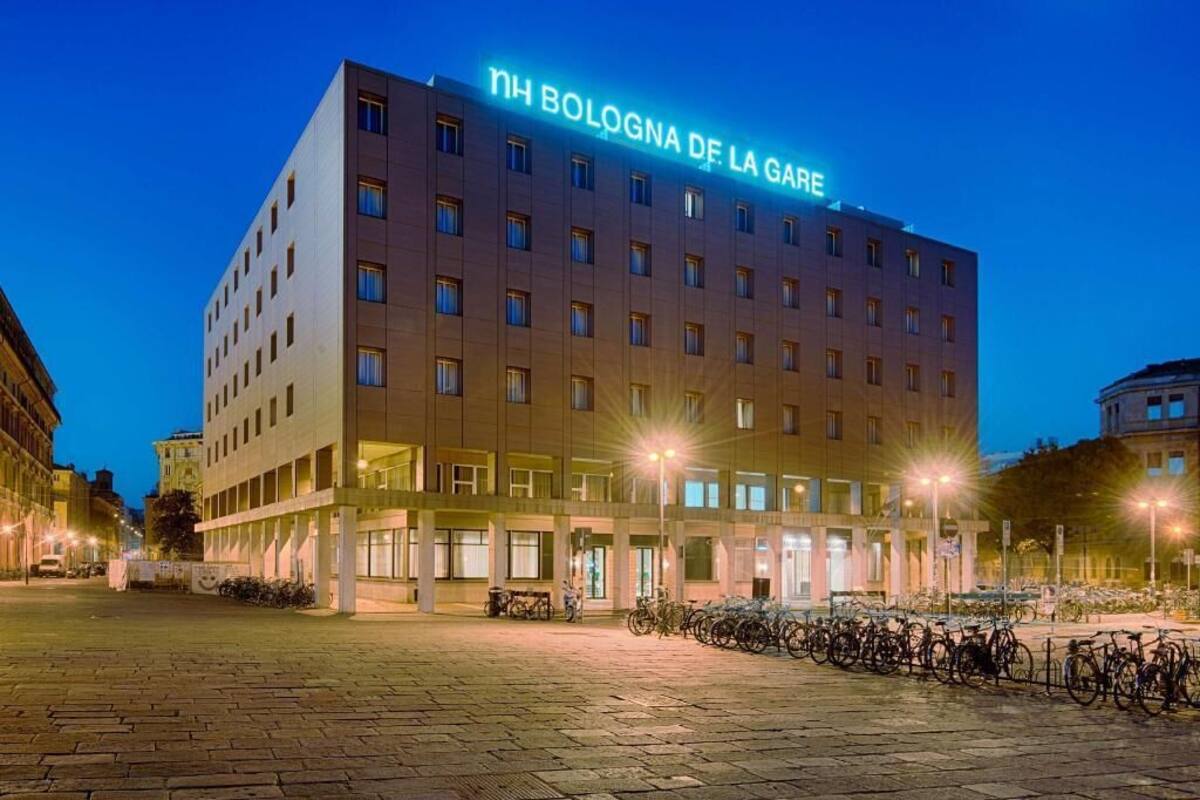 A hotel with a sign reading 'NH Bolognà DF. La Gàre,' featuring a modern facade, multiple floors, and a parking area with bicycles.
