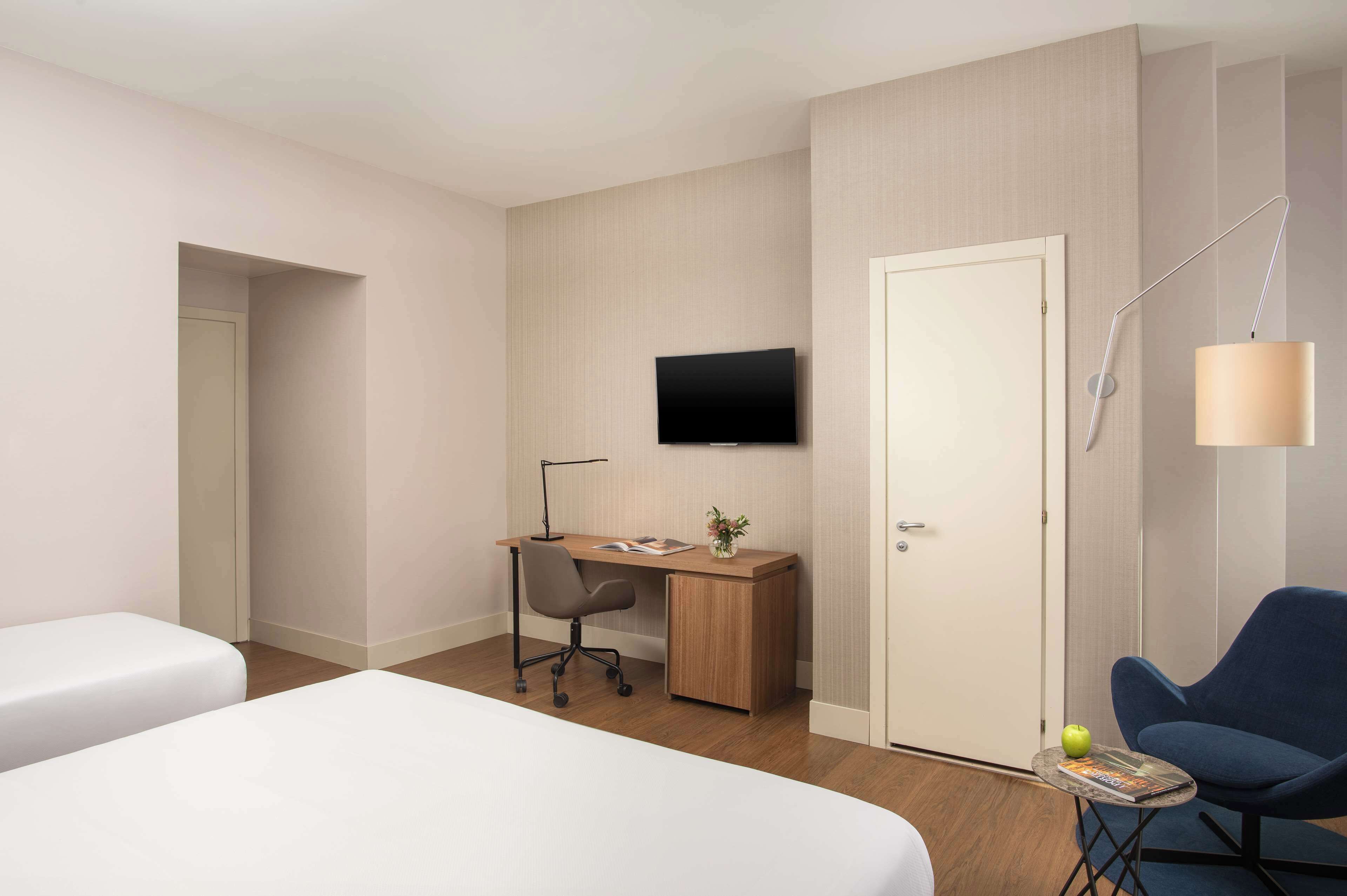 Quarto de hotel com duas camas, uma escrivaninha com cadeira, televisão, candeeiro e poltrona.