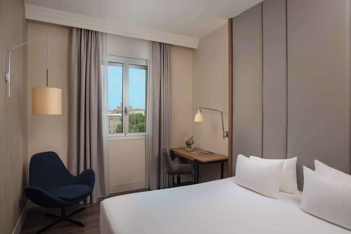 A hotel room with a bed, a desk, a chair, a lamp, and a view of buildings through the window.