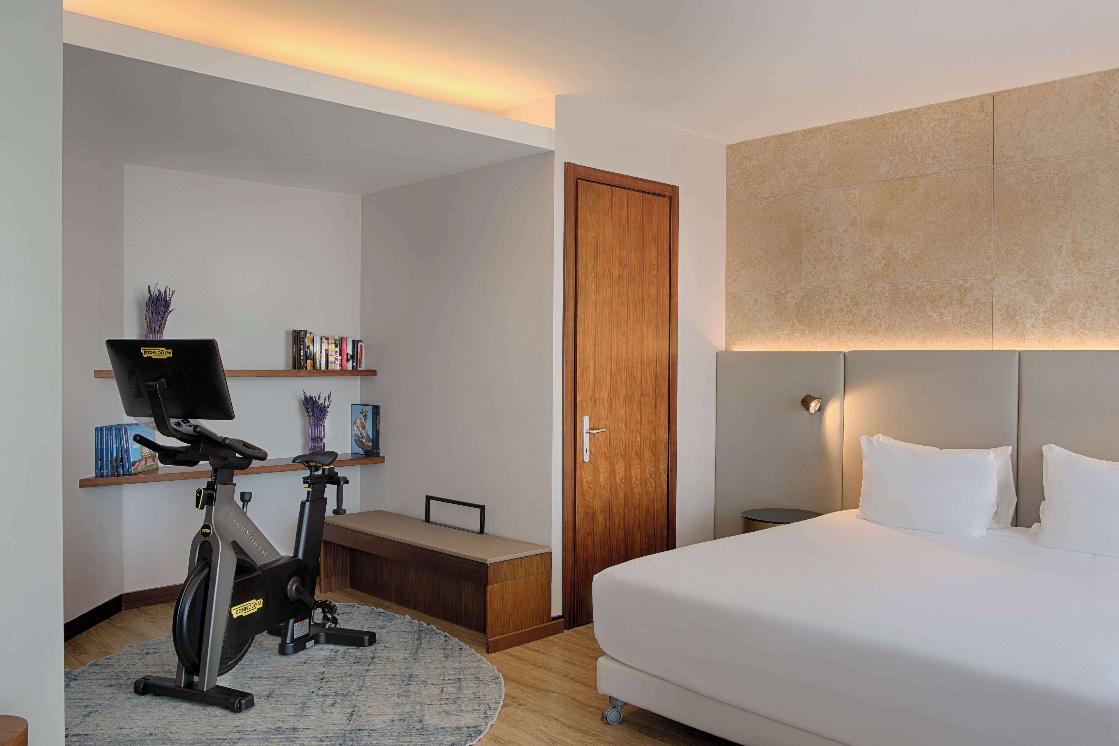 Habitación de hotel con cama, escritorio con computadora, pupitre con silla, caminadora y estantería.