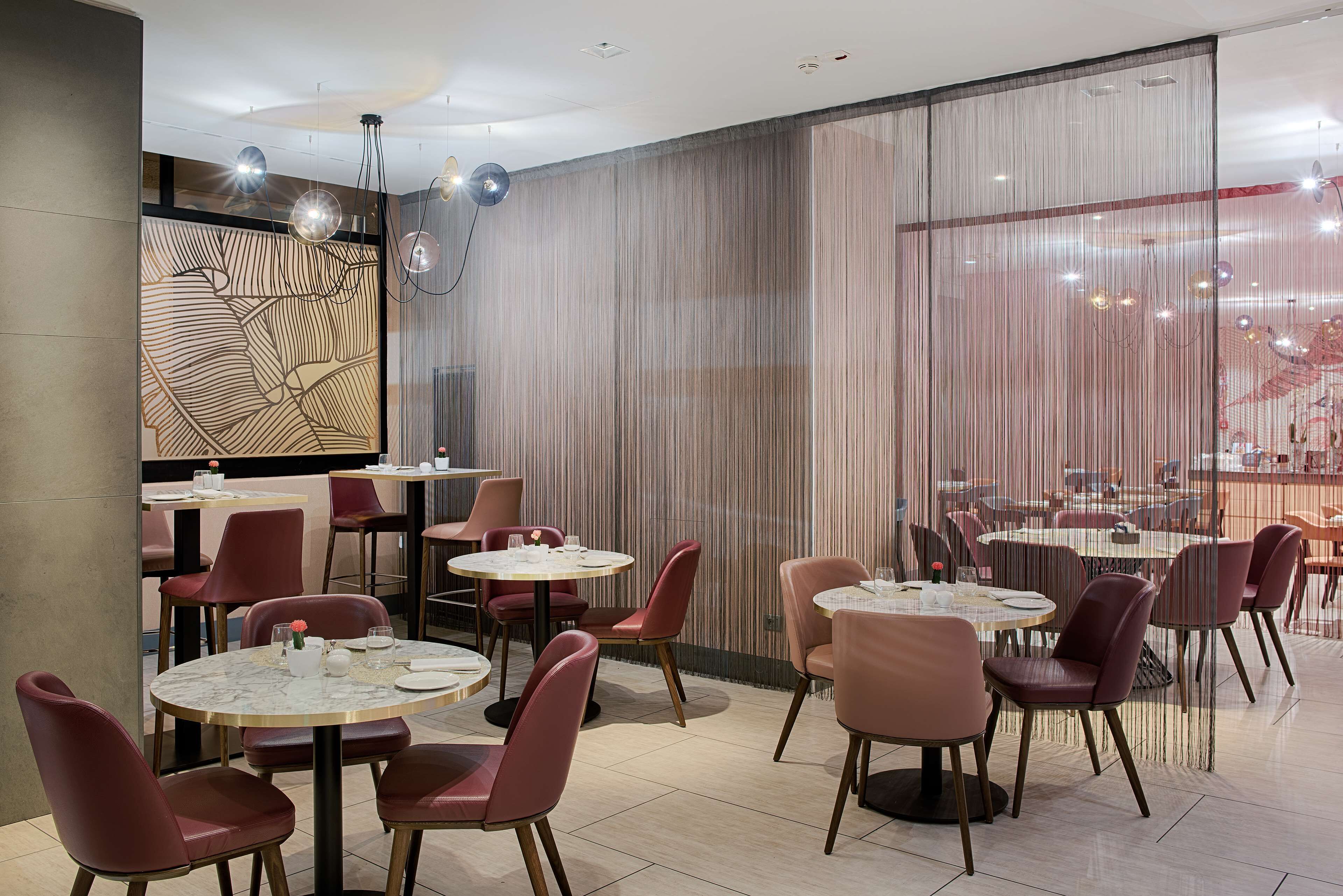 A modern restaurant interior with round tables and cushioned chairs, a decorative wall with a geometric pattern, and a large window with a view.