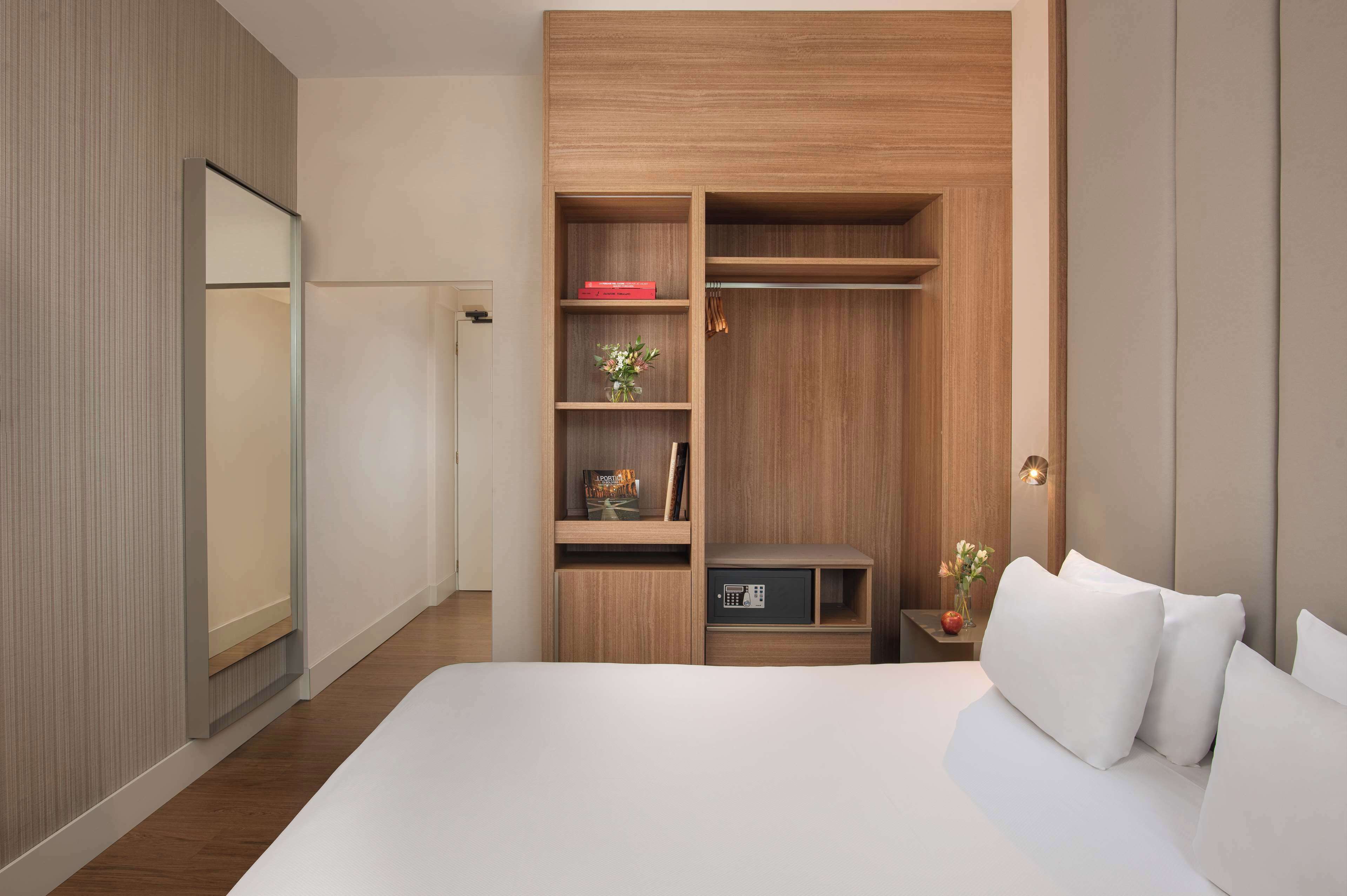 Ein modernes Hotelzimmer mit einem großen Bett, einem Holzschrank, einem kleinen Nachttisch mit einer Lampe und einem Beistelltisch mit Blumen.