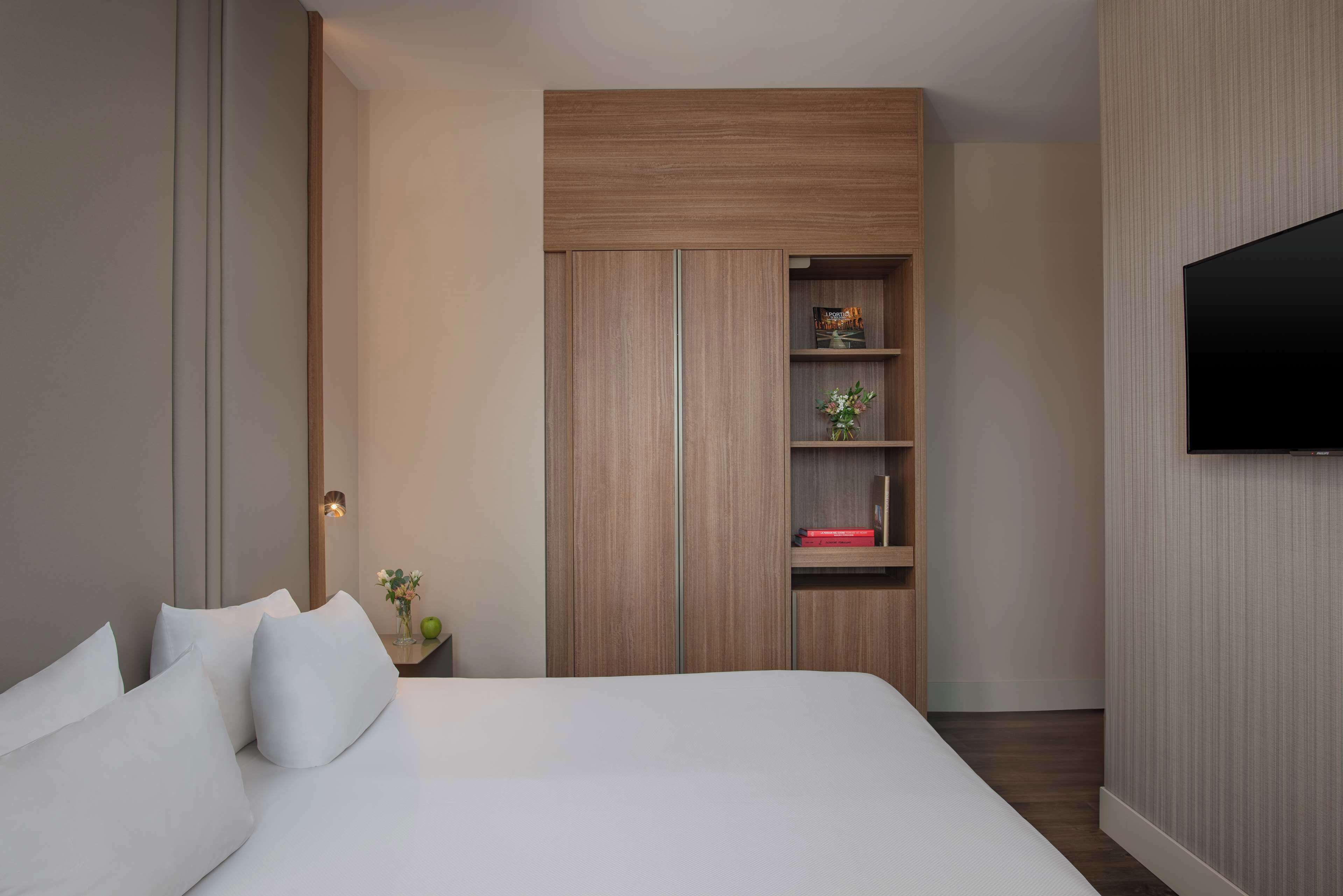 A modern hotel room with a large bed, a wooden wardrobe with shelves, a small table with a vase, and a flat-screen TV mounted on the wall.