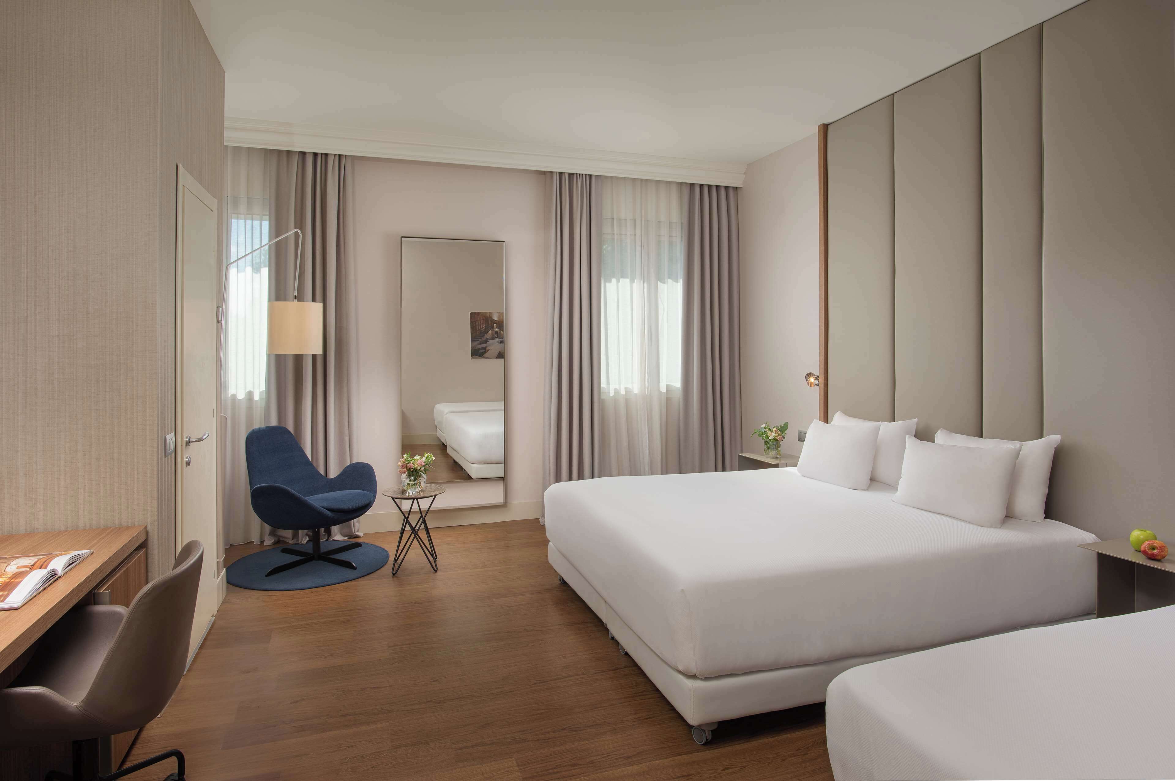 Habitación de hotel con dos camas, un escritorio, una silla, una mesita con lámpara y un espejo.
