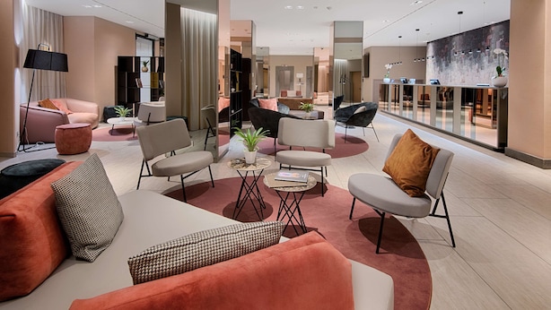 Un moderno vestíbulo de hotel con un área de estar que incluye sofás y sillones, una mesa de centro con libros, y un espejo grande que refleja el espacio.