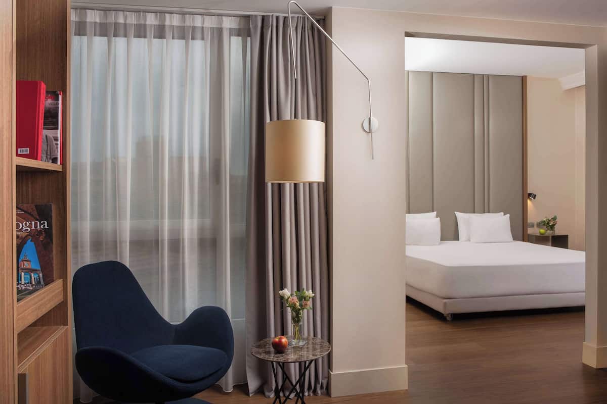 A hotel room with a bed, a chair, a small table with a vase, and a bookshelf.