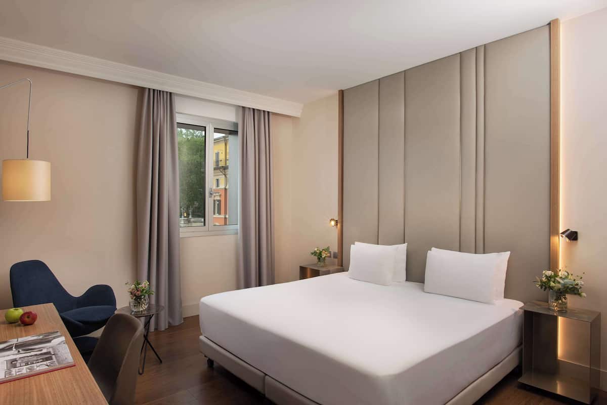 A hotel room with a large bed, a desk with a chair, a lamp, and a window with curtains.