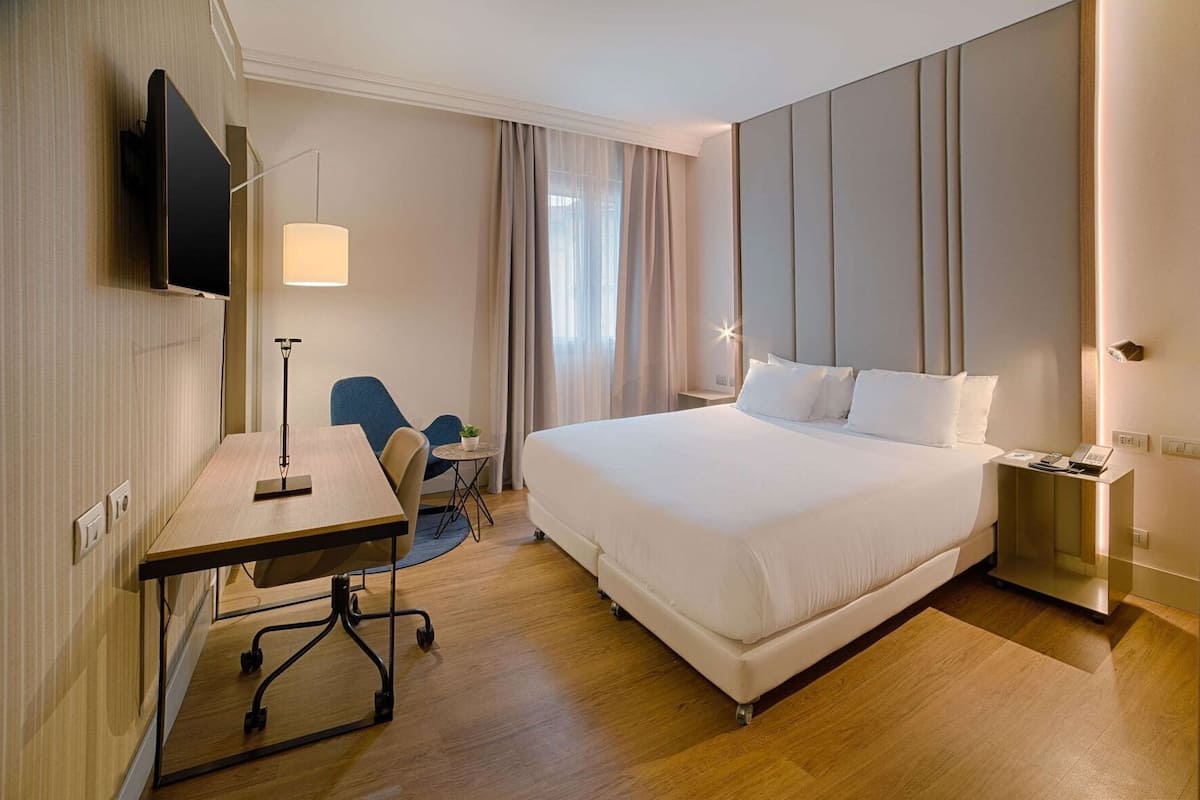 A hotel room with a bed, desk, chair, lamp, and bedside table.