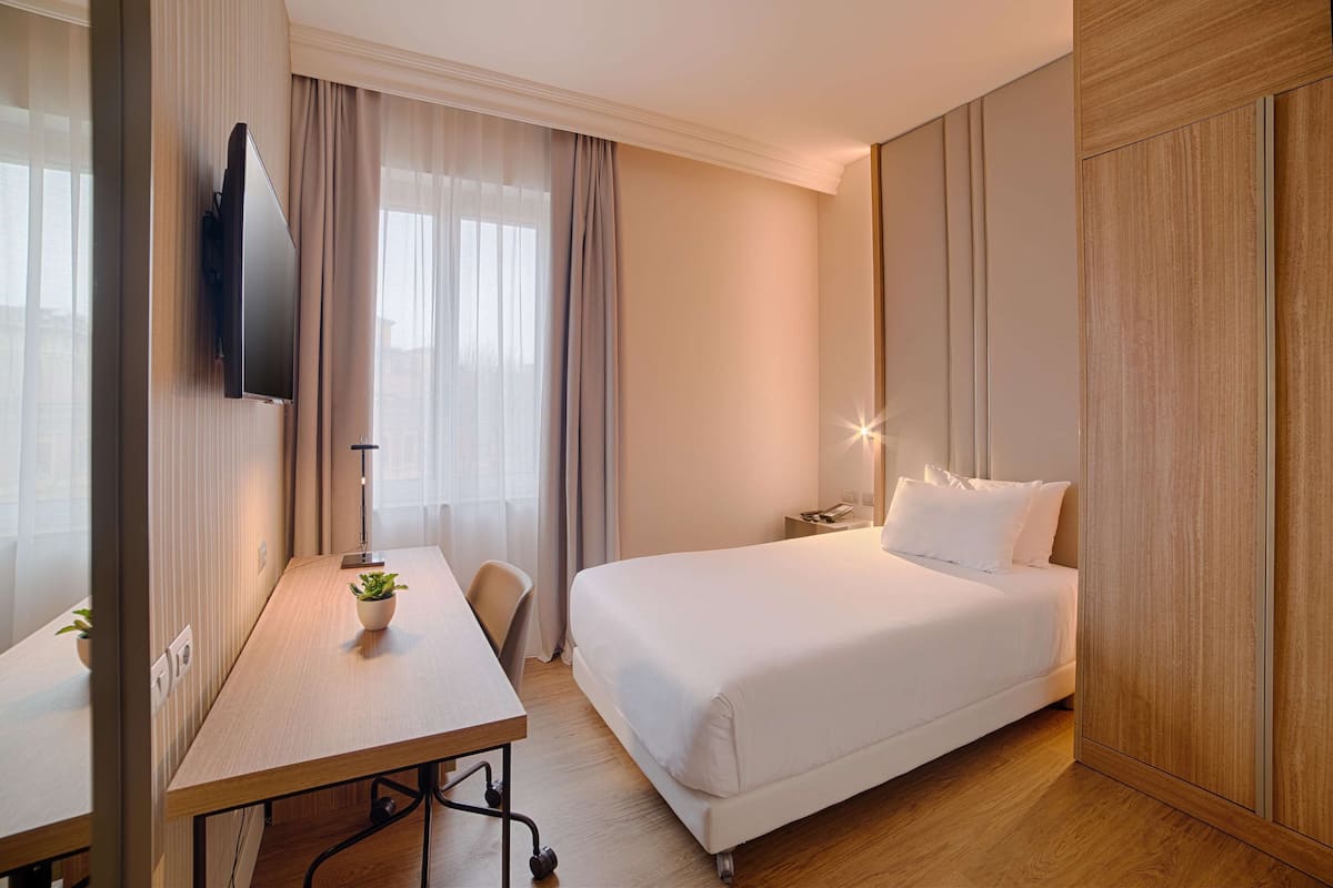 A hotel room with a bed, a desk, a TV mounted on the wall, and a nightstand with a lamp.