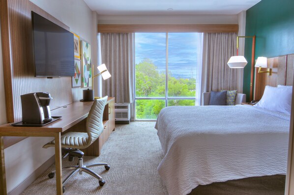 Habitación de hotel con cama, escritorio, silla, televisión y ventana grande con cortinas.