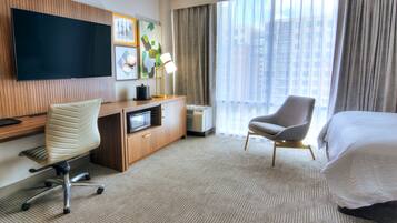 A hotel room with a large flat-screen TV, a desk with a chair, a bed, and a window with curtains.