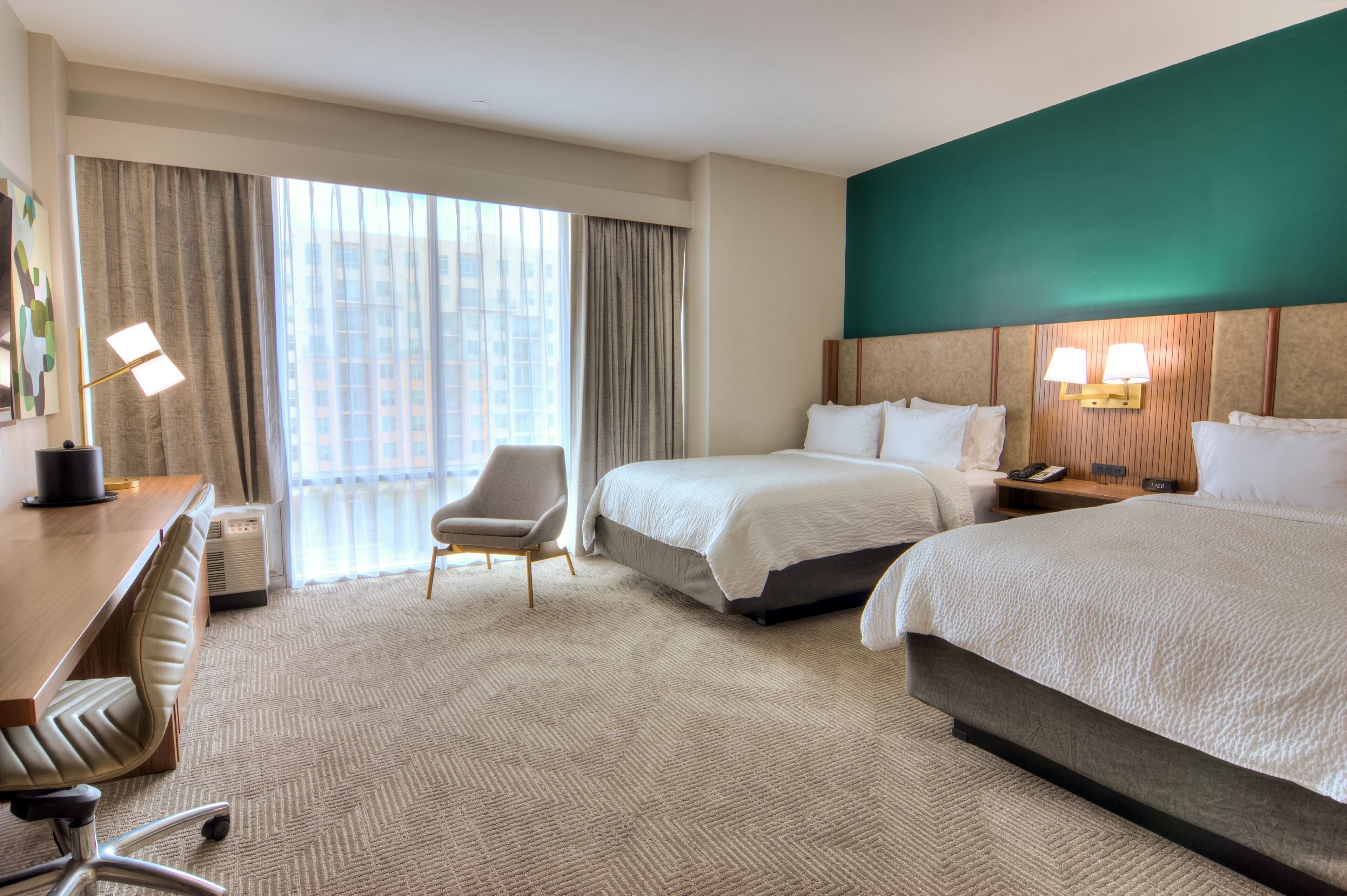 Habitación de hotel con dos camas, un escritorio, una silla, un ventanal con cortinas y una pared de acento verde.