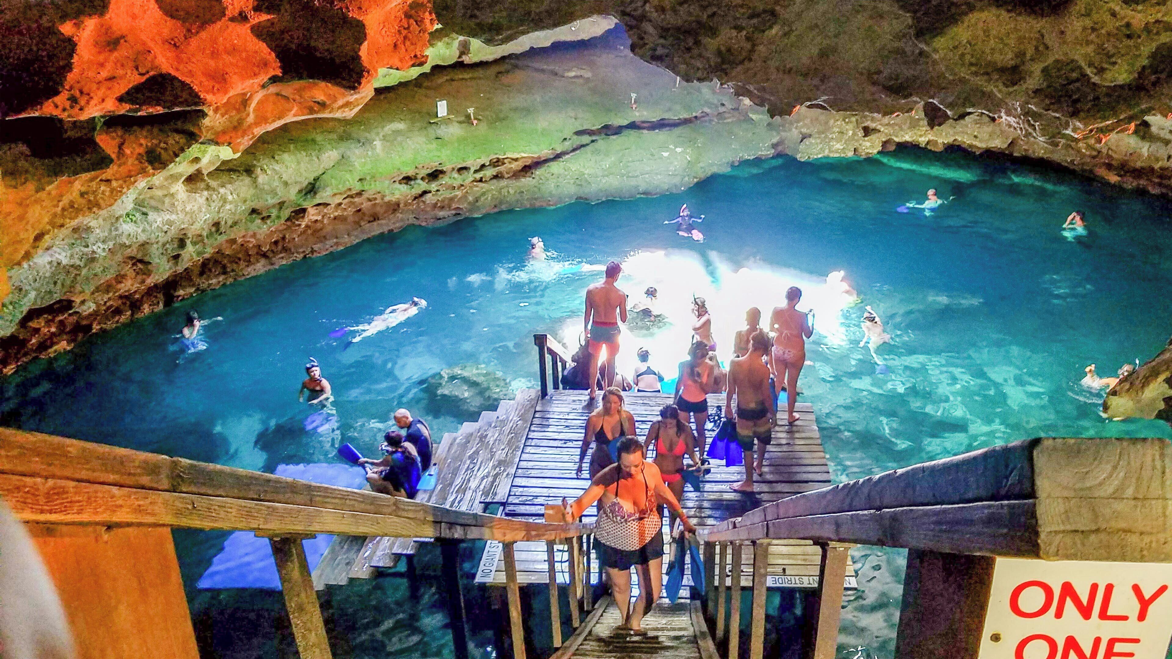 Un espace piscine avec une eau claire, des marches en bois menant à la piscine et un panneau indiquant un point d’entrée unique.