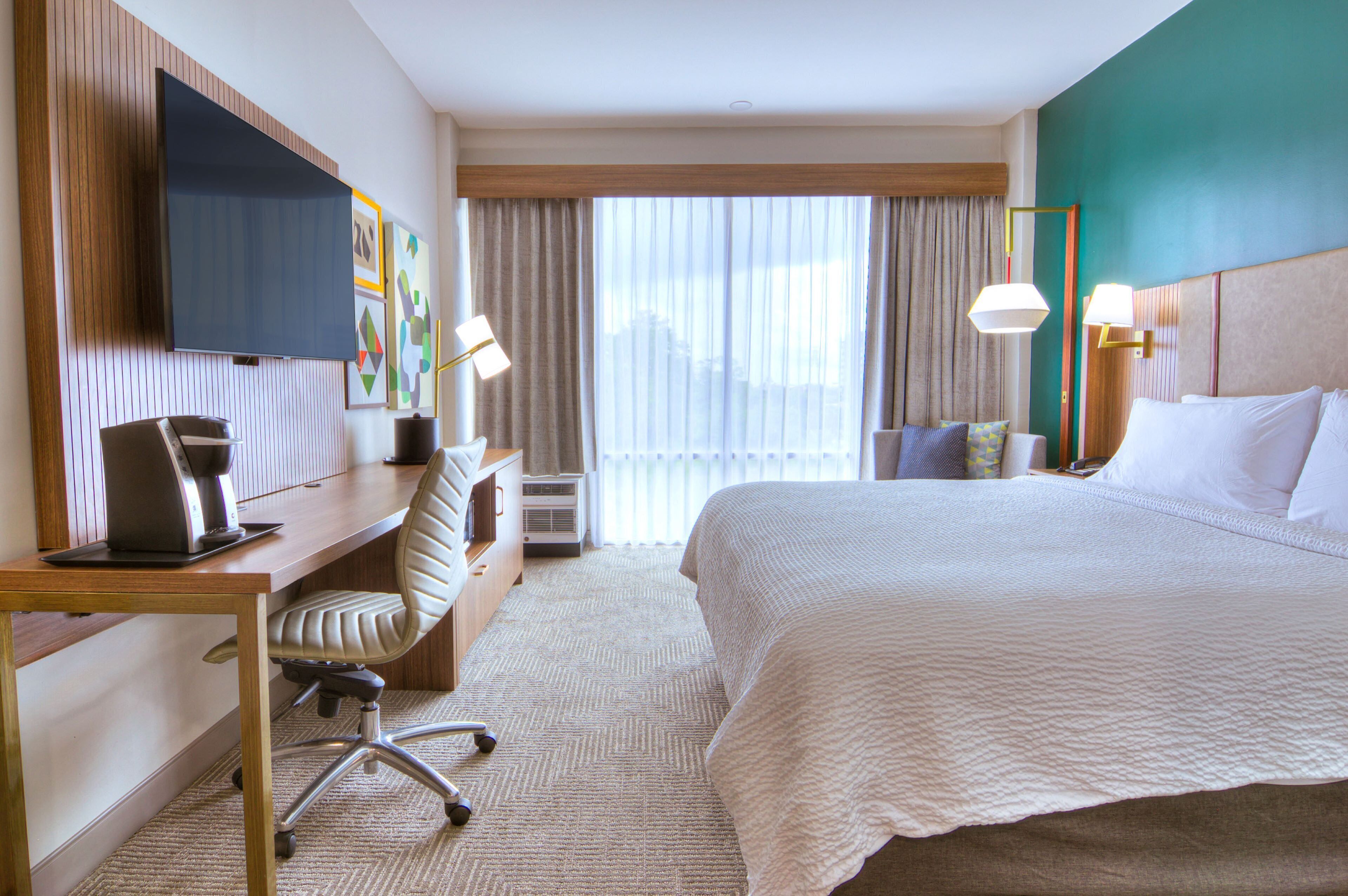A hotel room with a bed, desk, chair, TV, and large windows.