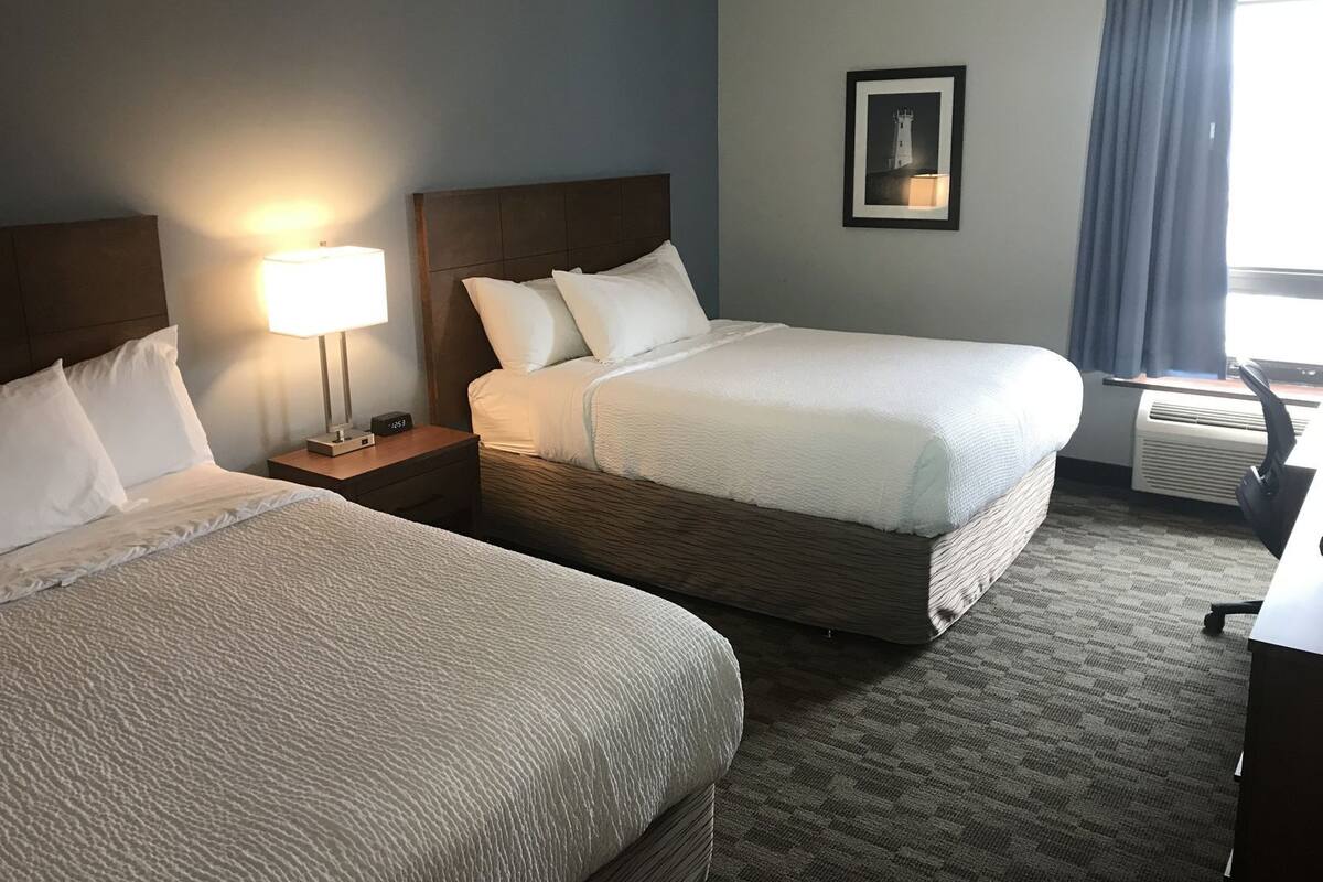 Une chambre d’hôtel avec deux lits, une table de chevet avec une lampe, un tableau encadré au mur et un appareil de climatisation.