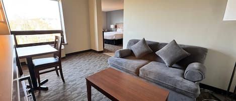 A hotel room with a grey sofa, a wooden coffee table, a desk with a chair, and a large window.