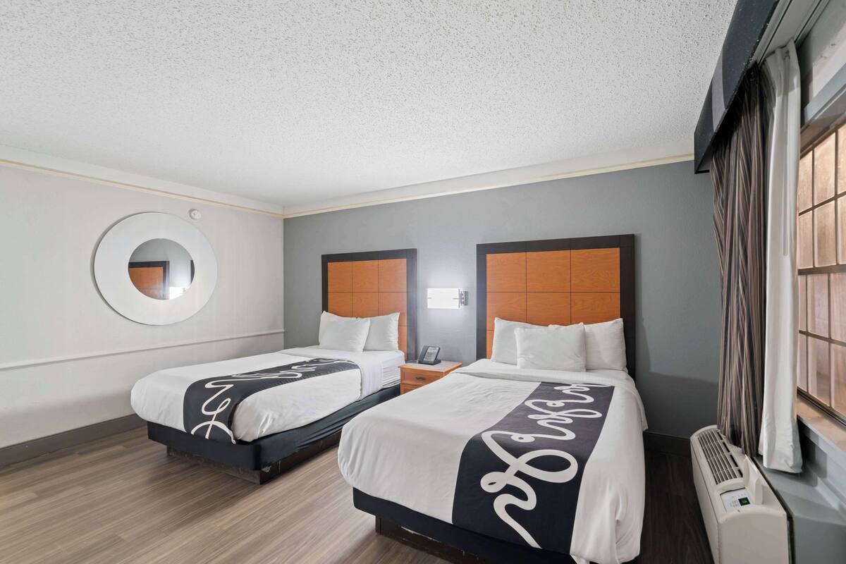 Une chambre d’hôtel avec deux lits, un miroir rond, une table de chevet avec un téléphone et une fenêtre avec des rideaux.