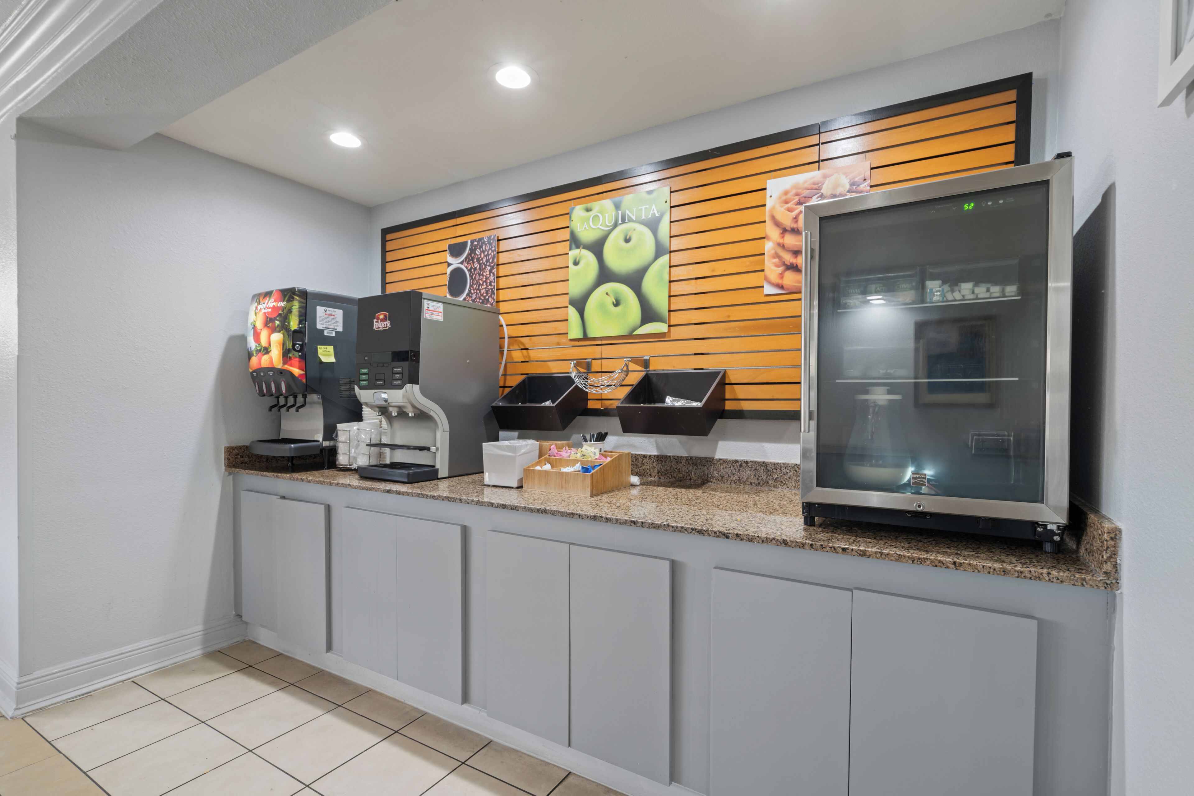 A modern kitchen area with a coffee machine, a refrigerator, and a wooden wall with a fruit-themed poster.