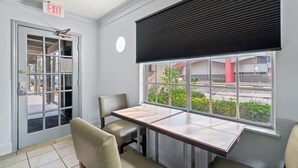 Een kamer met een tafel en twee stoelen, een raam met zonwering, een vluchtroutebord en een deur met uitzicht op buiten.