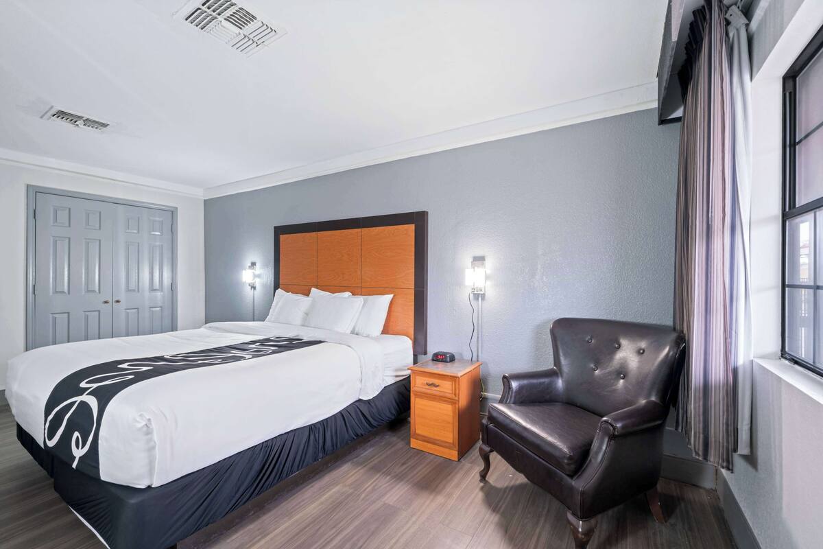 Une chambre d’hôtel avec un lit, une table de chevet en bois, un fauteuil en cuir et une fenêtre avec des rideaux.