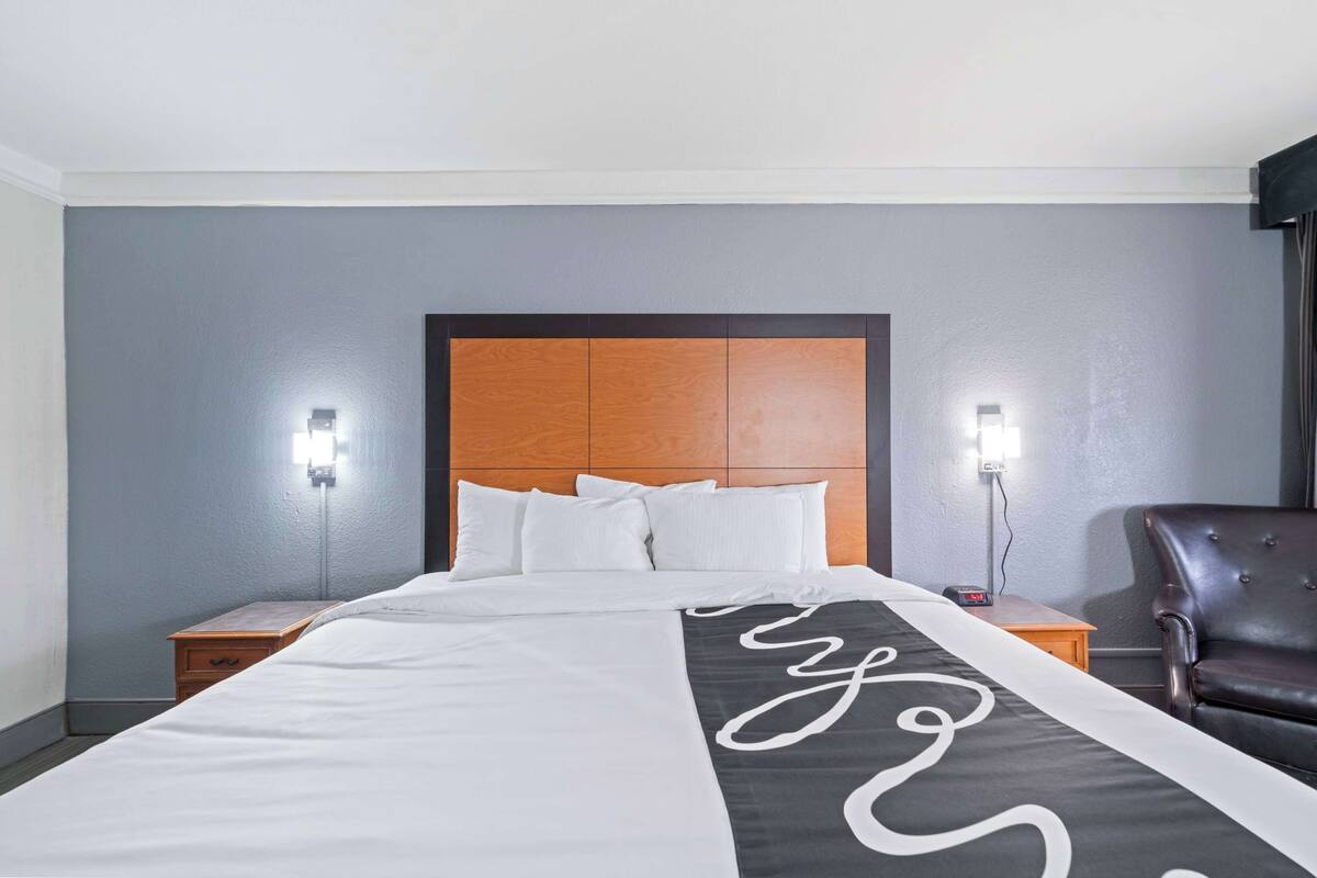 Une chambre d’hôtel avec un grand lit, deux tables de chevet, une tête de lit en bois, un fauteuil en cuir noir et deux lampes fixées au mur.