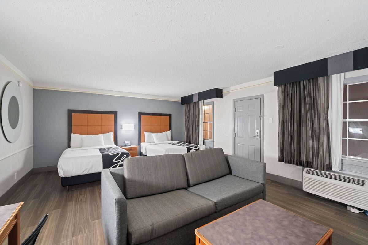 Une chambre d’hôtel avec un canapé gris, deux lits simples dotés de têtes de lit en bois, une petite table et une grande fenêtre avec des rideaux.