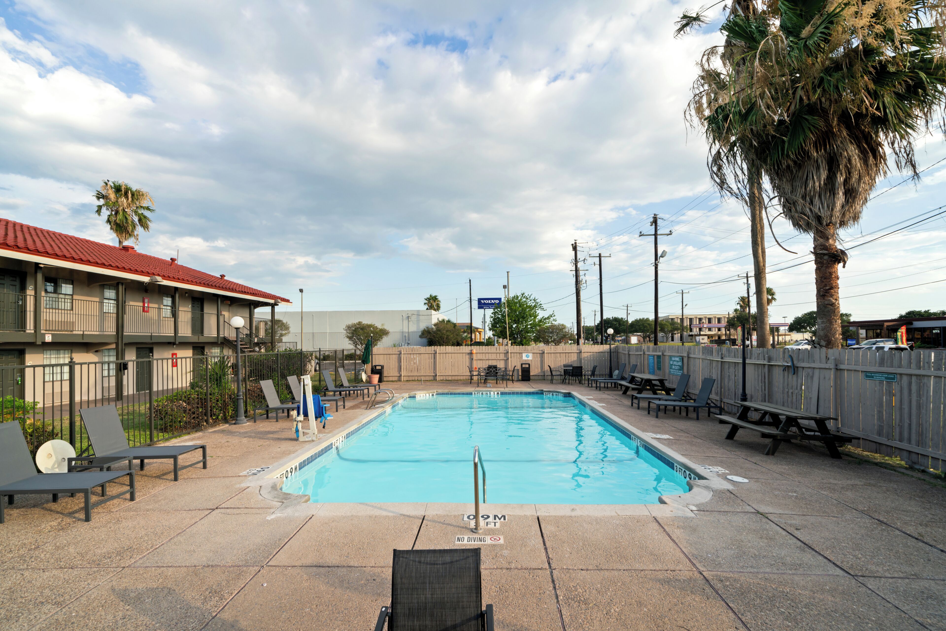 A rectangular swimming pool with a no diving sign, surrounded by lounge chairs and picnic tables, with a building and palm trees in the background.