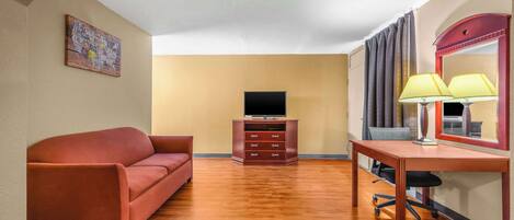 A hotel room with a wooden floor, a red sofa, a desk with a lamp, a television, and a dresser.