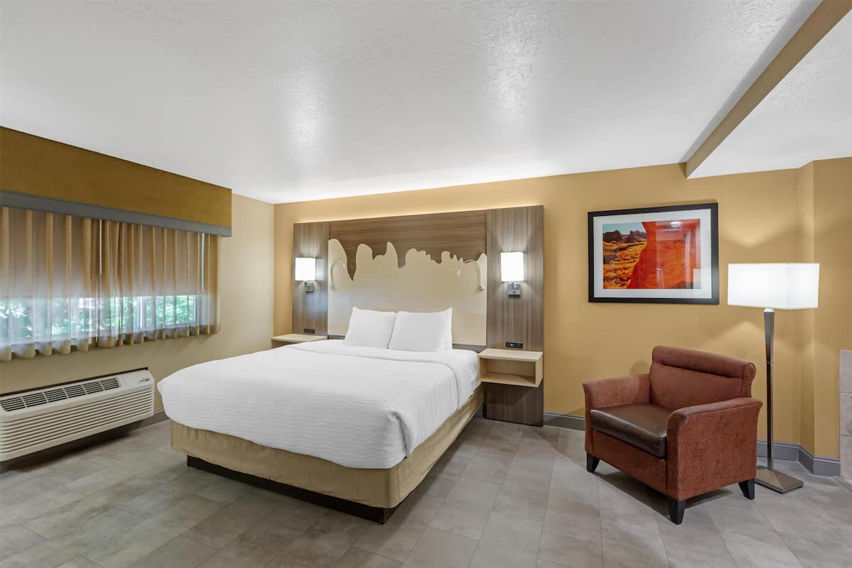 A hotel room with a large bed, a chair, a nightstand, a lamp, and a painting on the wall.