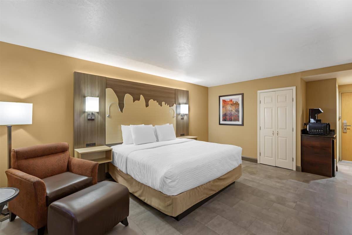 A hotel room with a large bed, a brown armchair, a small table, a desk with a computer, and a door leading to another room.