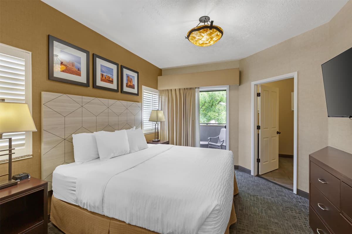 A hotel room with a large bed, two bedside lamps, a ceiling light fixture, a flat-screen TV, and a door leading to a balcony.