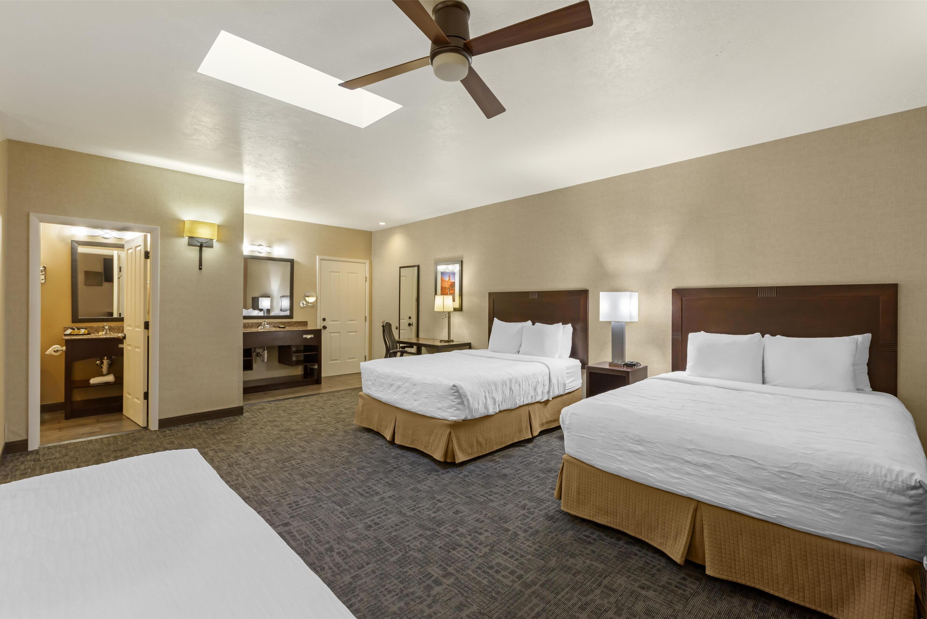 A hotel room with two beds, a ceiling fan, a desk, a chair, a mirror, and a bathroom with a sink and mirror.